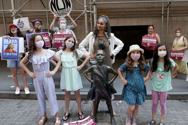 Maya Wiley stands with her hands on her hips behind the statue of the "Fearless Girl" which is a statue of a girl looking defiant with her hands on her hips, with little girls also mimicking the pose